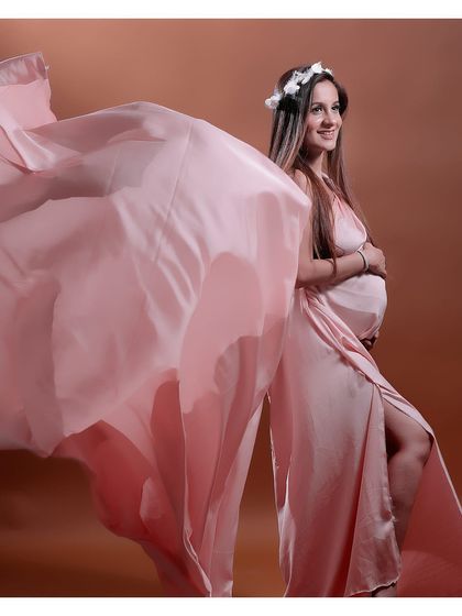 A whimsical shot of a mother-to-be in a flowing pink satin gown with a floral crown. The fabric is tossed to create a beautiful sense of motion.