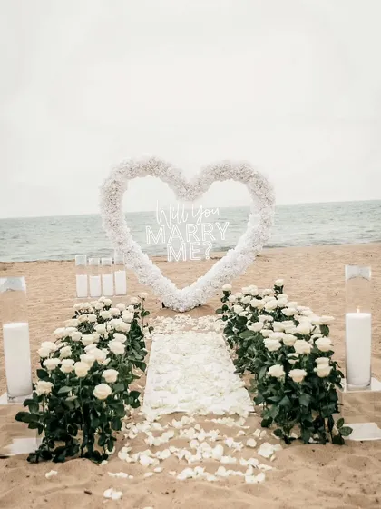 I transform beautiful locations like this beach into a private paradise for your proposal. This elegant setup features a white floral heart, a petal-strewn aisle, and candlelight to create a truly romantic engagement setting.