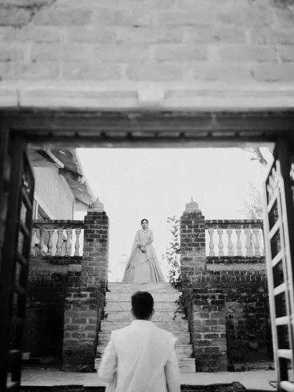 A dramatic black and white 'first look' style shot. The groom waits at the bottom of the stairs as his bride appears, a moment full of anticipation and romance, framed beautifully by the doorway.