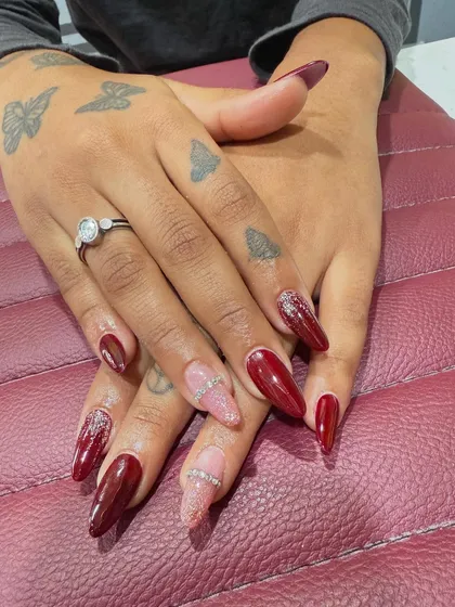A classic look with a touch of glamour. This set features long, almond-shaped nail extensions in a deep, glossy red, with accent nails in soft pink glitter and a delicate rhinestone band for extra sparkle. The client's hand tattoos show how well body art and nail art can complement each other.