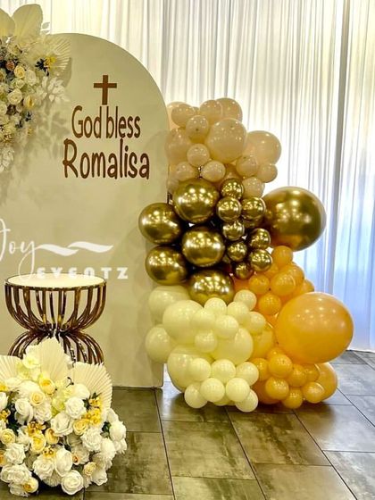 A pastel-themed decoration for a First Communion, using a soft yellow and gold color palette. The design includes a balloon garland, floral arrangements, and an elegant custom backdrop.