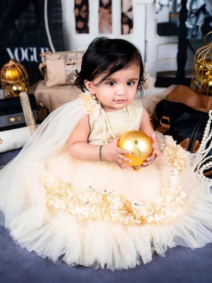 A baby girl in a beautiful cream dress holds a golden ball, looking serene and poised in her stylish setup.