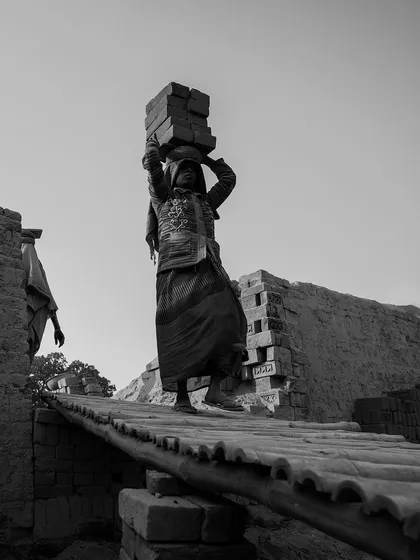 A worker carefully balances a load of bricks on her head while walking across a narrow makeshift bridge, a black and white photo highlighting her skill and focus.