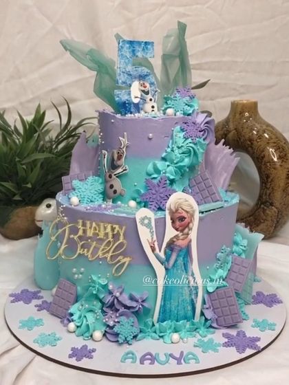 A magical two-tier Frozen themed cake for a 5th birthday. This design features Elsa, Olaf, and beautiful snowflake details in shades of purple and teal, all created with whipped cream.