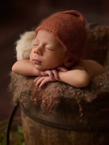 A newborn in a rust-colored knitted hat sleeps in a dark wooden bucket lined with soft, textured wool.