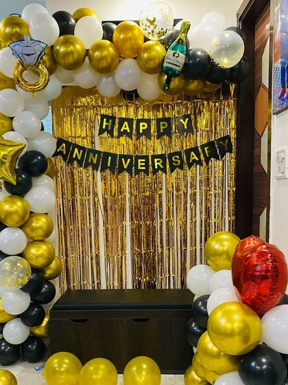 A classic anniversary decoration for your home. This setup includes a balloon arch in black, white, and gold over a foil curtain, with a "Happy Anniversary" banner.