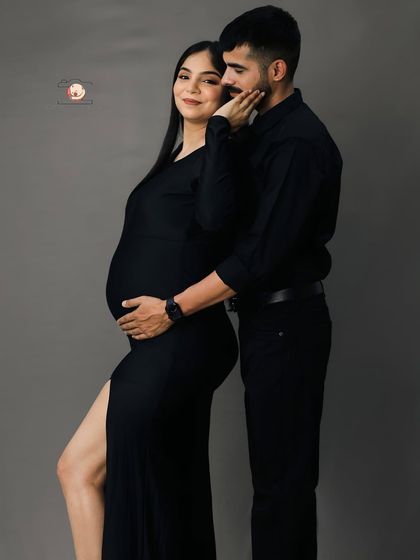 A chic and modern couple's portrait. The partner stands behind, embracing his wife, who is dressed in a stylish black dress with a side slit.