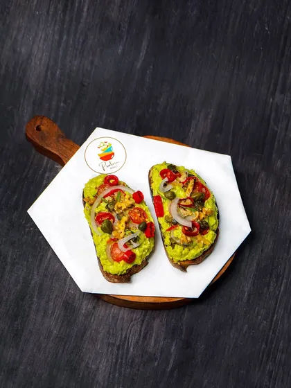 Avocado toast topped with onions, tomatoes, and capers, shot from above on a dark wooden surface. The moody lighting highlights the vibrant green of the avocado.