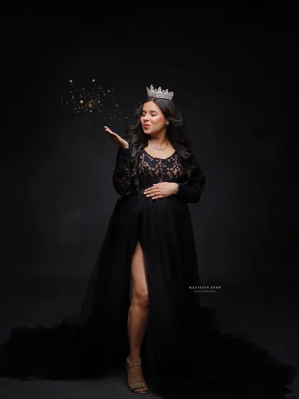 A magical portrait of a queen. The mother-to-be wears a crown and a black lace gown, blowing a puff of gold glitter into the air.