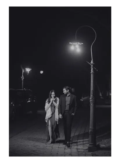 A romantic black and white photo of the couple walking down a street at night, illuminated by a single lamppost. This image has a classic, old-world cinematic charm.