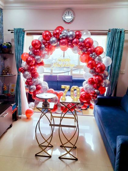 A festive 70th birthday anniversary celebration at home. A circular balloon arch in red and silver frames the living space, with a light-up marquee '70' as the centerpiece.
