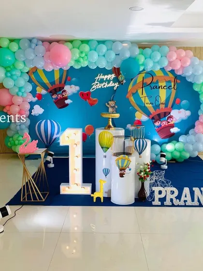 This hot air balloon theme is perfect for a 'flying high' first birthday party. The wide backdrop creates an immersive sky scene, complemented by a pastel balloon garland, marquee number, and themed props.