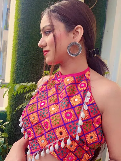 A close-up of a vibrant pink and orange halter neck blouse with mirror work and shell tassels, a modern twist on Garba wear.
