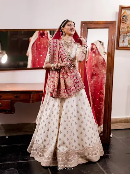 A bride in a beautiful white and red lehenga smiles at her reflection in an antique mirror, a perfect moment of pre-ceremony joy.
