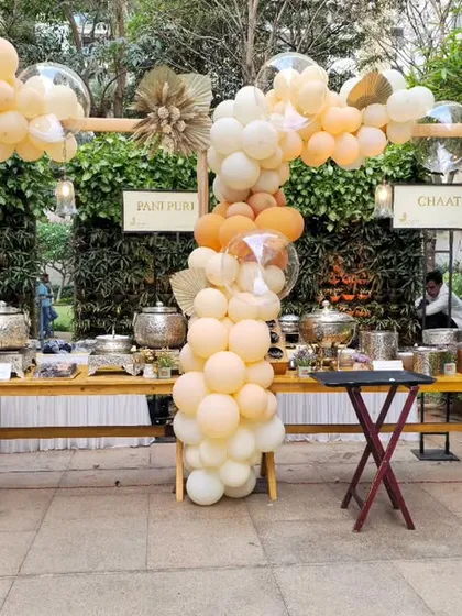 A stylishly decorated food station at an outdoor event. The buffet tables are framed by two balloon columns in neutral, earthy tones.