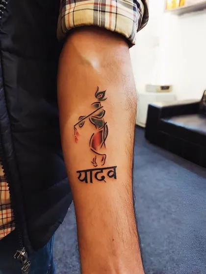 A minimalist depiction of Lord Krishna playing the flute, paired with the client's family name 'Yadav'. This tattoo blends personal identity with spiritual devotion.