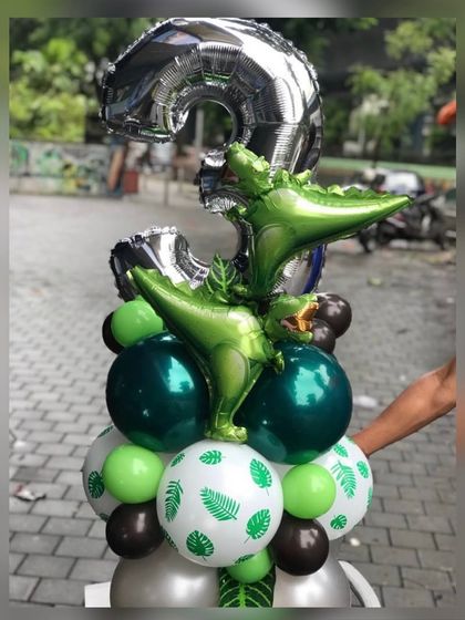 A close-up of a dinosaur-themed balloon bouquet for a 3rd birthday. This arrangement features a large silver number "3" foil, dinosaur foils, and leaf-printed balloons in a green and silver color scheme.