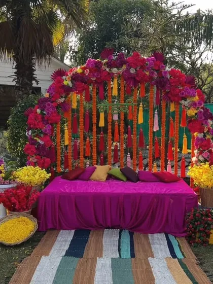 A colorful and festive outdoor setup for a Haldi or Mehandi function. This decor uses a bright pink and orange floral arch with marigold tassels, a comfortable seating diwan, and baskets of fresh flowers, creating a perfect garden party vibe.