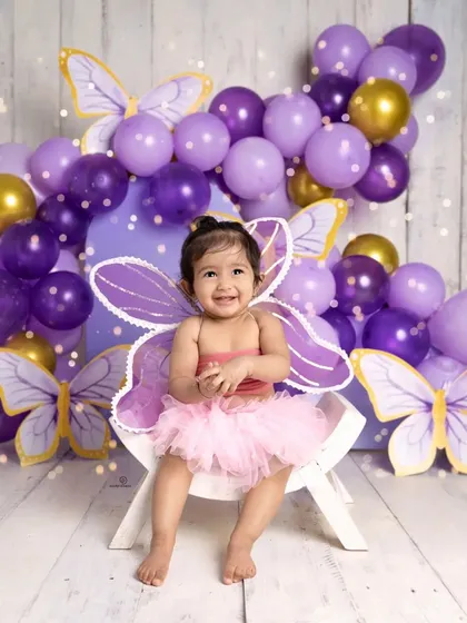 A happy toddler celebrates a milestone in a butterfly-themed photoshoot. Dressed in a pink tutu with purple wings, her joyful smile is captured against a backdrop of purple and gold balloons.