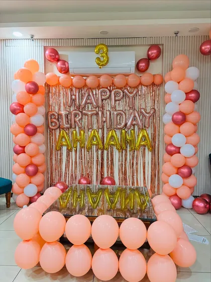 A beautiful 3rd birthday setup in peach, rose gold, and white. The decor includes balloon pillars and a tinsel backdrop with the birthday girl's name.