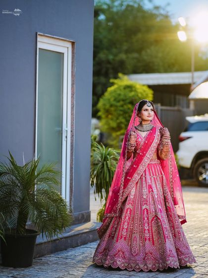 A stunning full-length portrait of the bride in her magnificent pink lehenga. The soft, natural light highlights the intricate details of her attire and her poised stance.