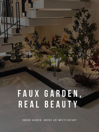 A diverse collection of artificial plants, including leafy greens and pops of red, creates a vibrant faux garden under a floating staircase. Set in a bed of white gravel with integrated lanterns, this design adds life and color to the space.