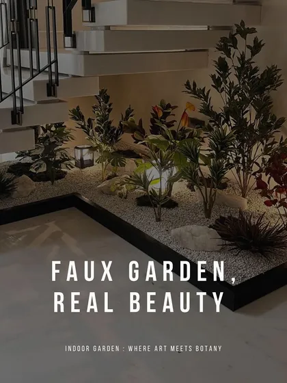 A diverse collection of artificial plants, including leafy greens and pops of red, creates a vibrant faux garden under a floating staircase. Set in a bed of white gravel with integrated lanterns, this design adds life and color to the space.