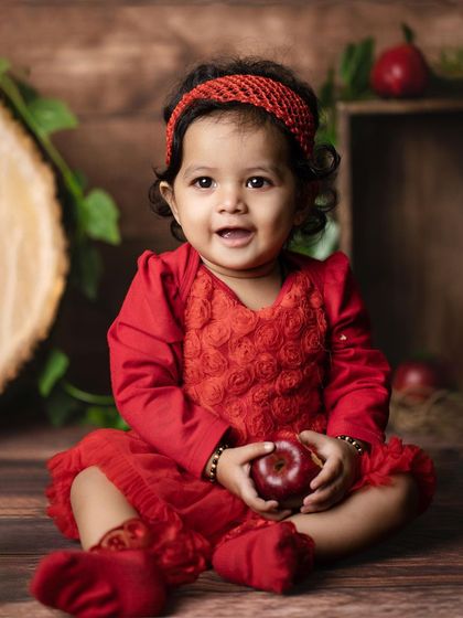 A happy baby girl in a bright red dress smiles for the camera, holding an apple in a rustic, themed studio setup. Her cheerful expression is absolutely infectious.