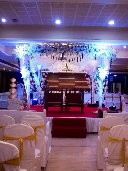 An elegant indoor mandap setup in a banquet hall. The design uses white drapes, a floral canopy, and coordinated chair covers to create a cohesive and beautiful ceremony space.