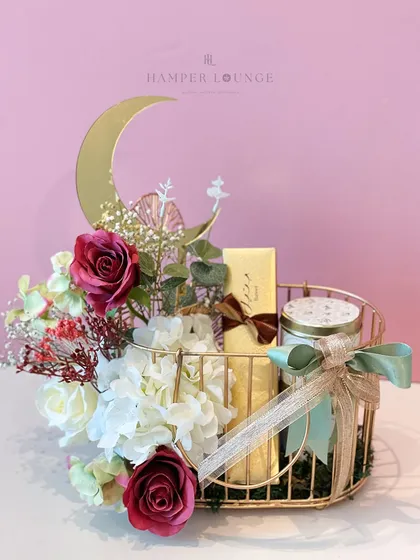 This Eid hamper features a golden metal basket adorned with a crescent moon. It's filled with a scented candle and other treats, surrounded by a delicate arrangement of red and white roses.