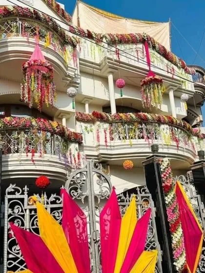 An incredibly grand and lavish house decoration with heavy floral garlands, chandeliers, and colorful drapes covering the entire facade. This is for clients who want an over-the-top celebration.