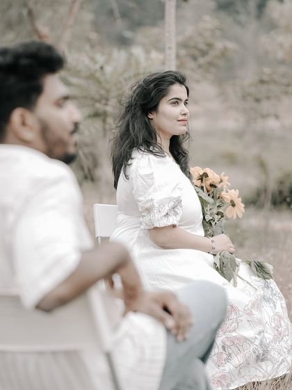 An artistic composition with the groom out of focus in the foreground, highlighting the bride sitting with a bouquet in a field.