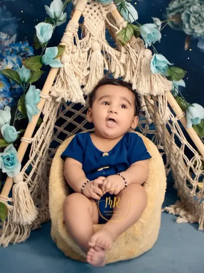 A baby boy sits in a small teepee decorated with blue flowers, looking up with an adorable, expressive face.