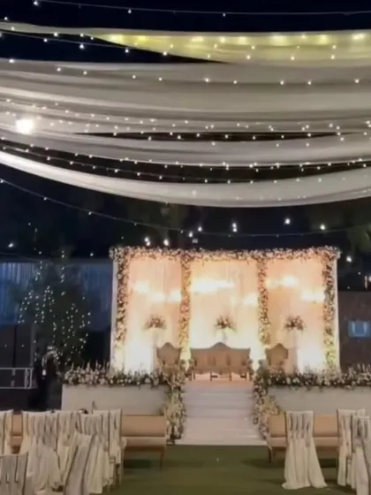 A wide view of an outdoor reception setup at night. The elegant white stage is complemented by a ceiling of fairy lights and drapes, creating a magical ambiance.