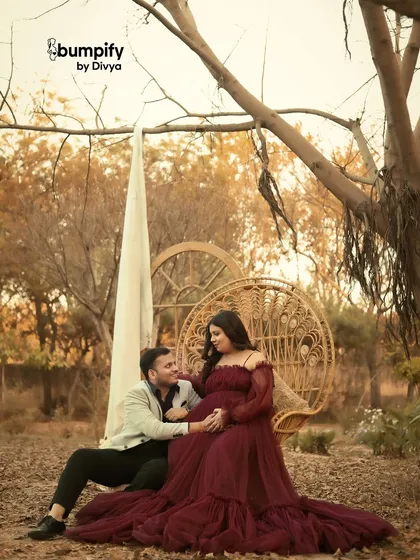 A romantic outdoor scene set against a warm, autumnal backdrop. The couple shares a moment on a wicker swing chair, with the mom-to-be in a rich maroon gown.