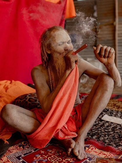 A Naga sadhu relaxes with his chillum at the Kumbh Mela. The red background and his orange robes create a warm, intimate portrait of a moment of rest.
