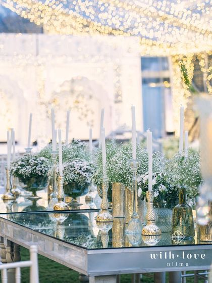 Tall candles and baby's breath arrangements on a mirror-topped table. The reflections create a dazzling effect, doubling the candlelight and the sparkle from the overhead fairy lights for a truly immersive experience.