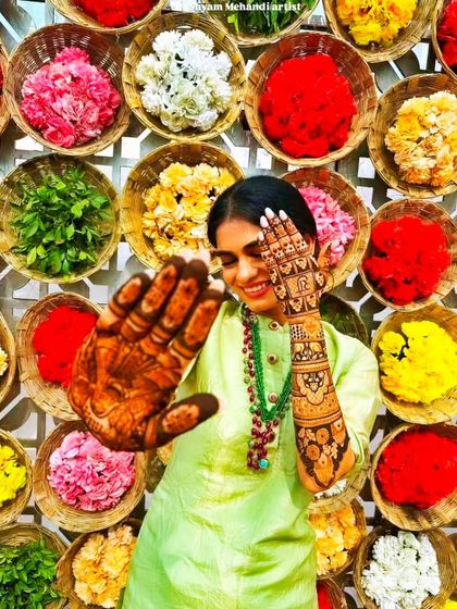 A creative and colorful shot of a client posing in front of a flower wall, her henna-adorned hand in the foreground.