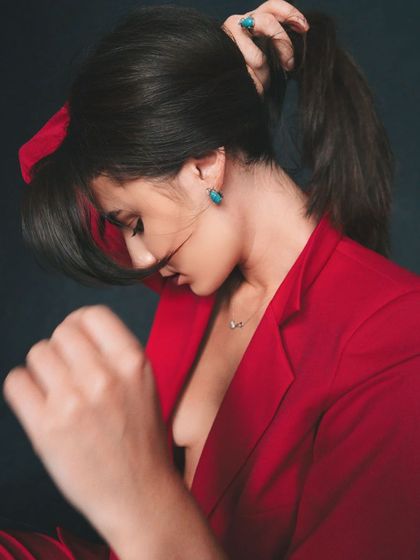 A candid-style portrait showing the model tying her hair back. The red blazer and dark background create a dramatic and stylish contrast.