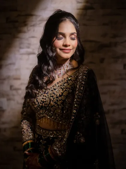 A dramatic and moody portrait. The lighting highlights the intricate embroidery on her dark lehenga and the soft glow of her makeup.
