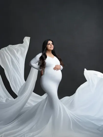 A powerful and statuesque portrait. The mother-to-be stands tall in a white gown, with the fabric flowing out like wings against a dark, dramatic backdrop.