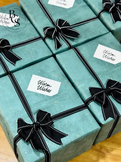 A set of corporate gifts wrapped in a textured teal paper. The look is completed with a stitched-edge black ribbon and a simple "Warm Wishes" tag, creating a polished and gender-neutral gifting option.