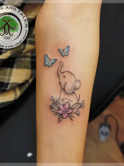 A delicate fine-line tattoo of a baby elephant with soft blue butterflies and a touch of pink in the flower. This shows our ability to create gentle, detailed pieces with subtle color accents.