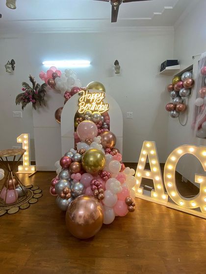 A stylish and modern first birthday decoration for a home party. This setup features a balloon cascade in rose gold, pink, and white, a 'Happy Birthday' neon sign, and large marquee letters.