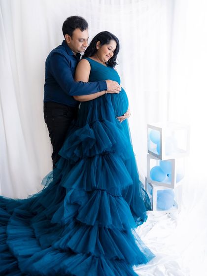 A sweet embrace from behind, with the couple looking down at the baby bump, surrounded by a soft white curtain and blue-themed props.