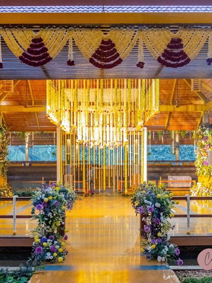 The entrance to a 'Sunrise Soirée' themed event, featuring floral pillars and a stunning hanging chandelier mandap in the background.