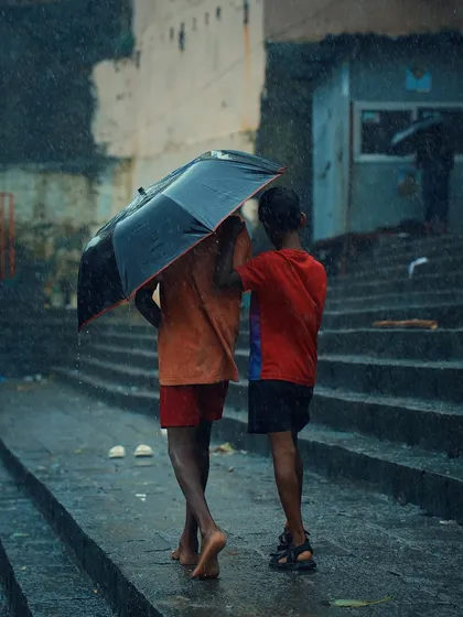 Two boys huddle together under a single umbrella, walking barefoot on the wet steps of a ghat. This is a heartwarming, candid photograph about friendship and finding shelter together in the rain.