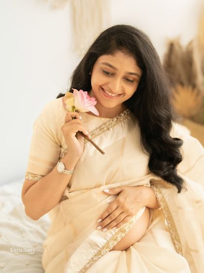 A sweet and delicate portrait of a mother-to-be in a cream saree. She is smiling as she holds a single pink lotus flower, captured in a soft and dreamy boho setting.