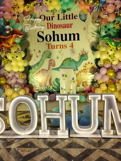 A shot focusing on the large, illuminated letters spelling "SOHUM" in front of the dinosaur-themed birthday backdrop.