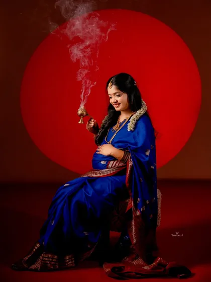 A striking portrait featuring a mom-to-be in a royal blue saree, holding a traditional incense burner. The bold red circular background adds a modern, graphic element to this cultural shot.